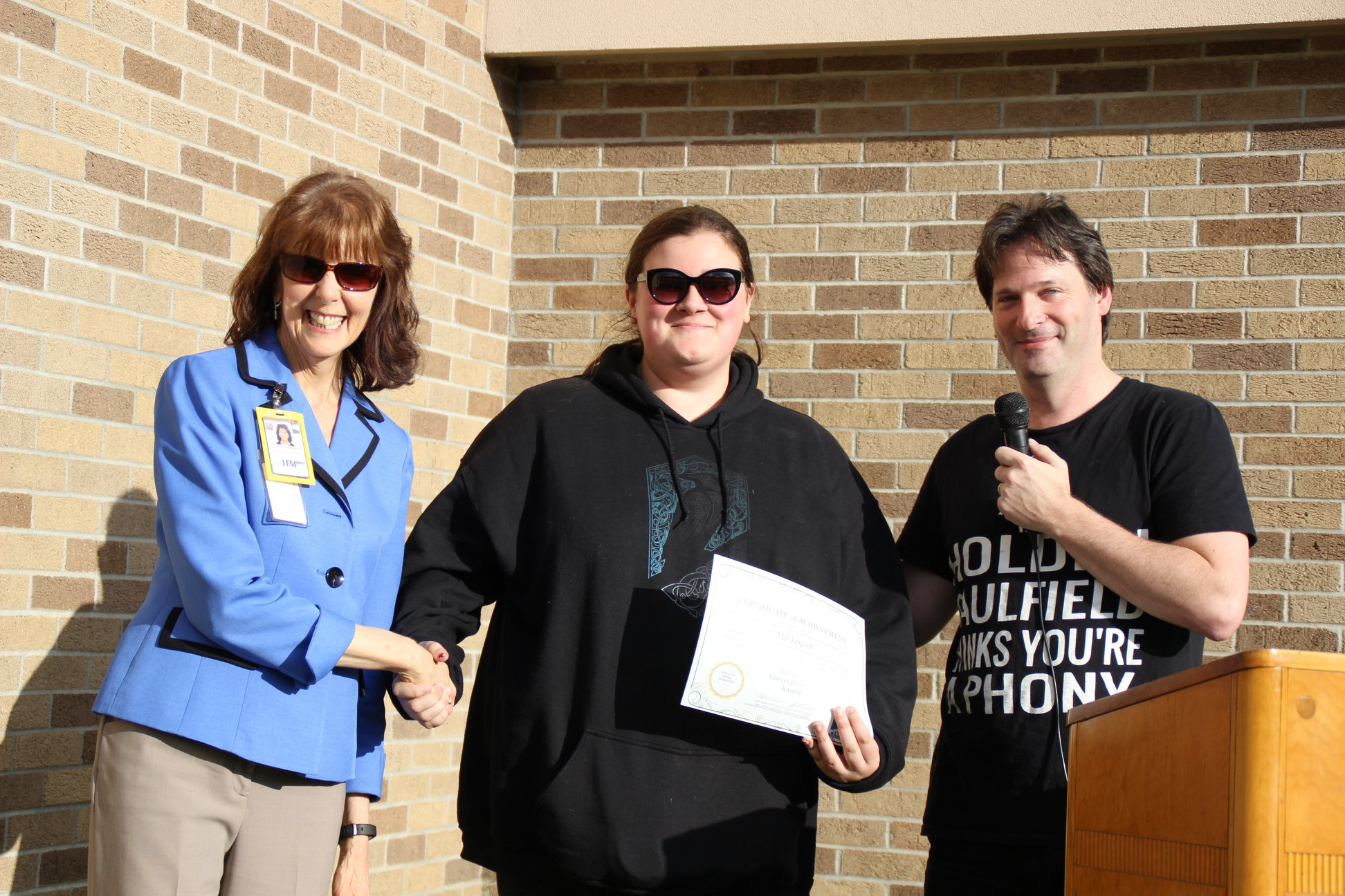 MJ Logan is pictured (center) receiving an award. She is shaking hands with a woman and is flanked on the other side by a man. They are all standing at a podium.