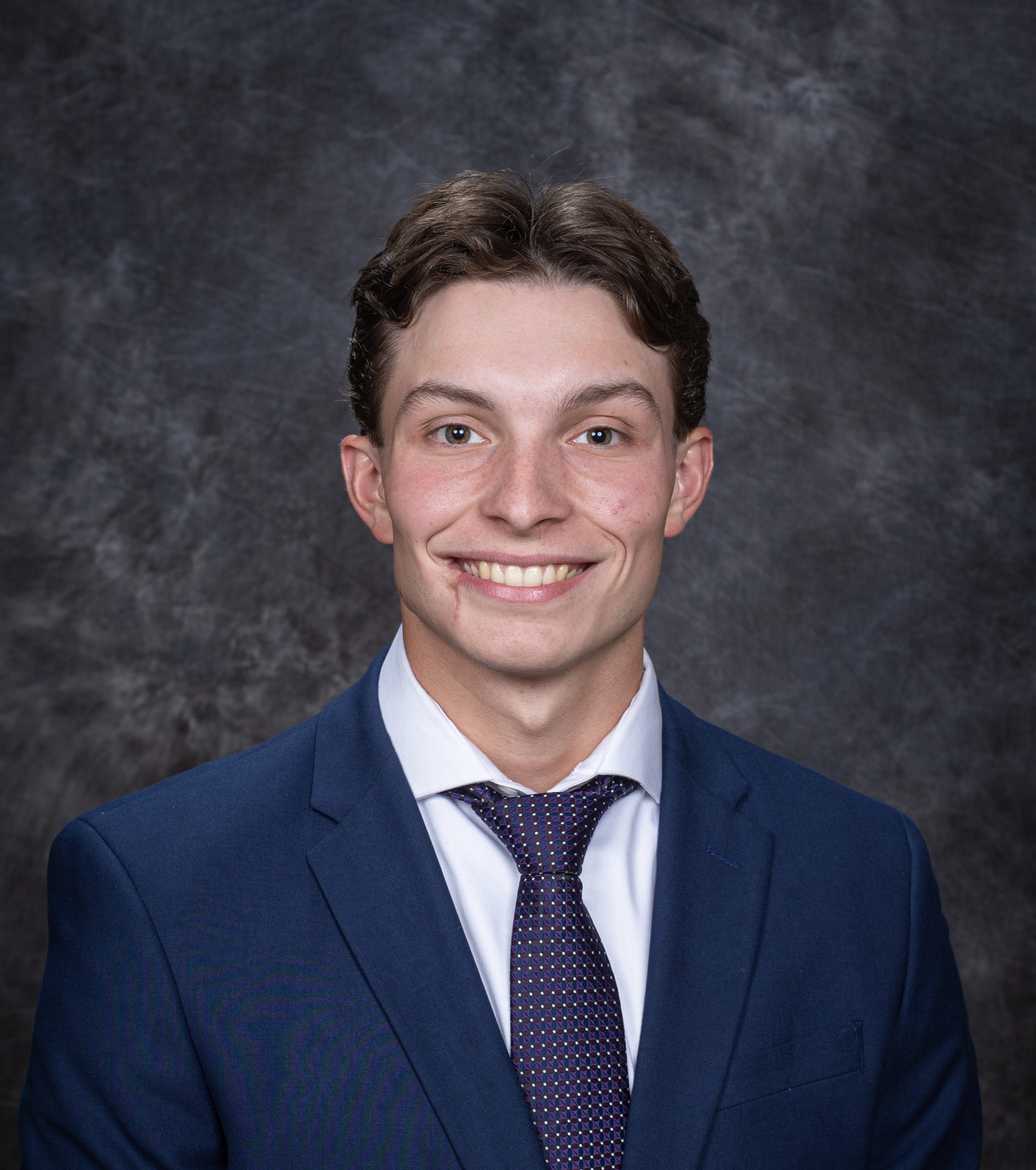 Headshot professional work photo of Frank Alvarez. He is wearing a white dress shirt with a navy sports coat and a blue and white tie. The background is gray.