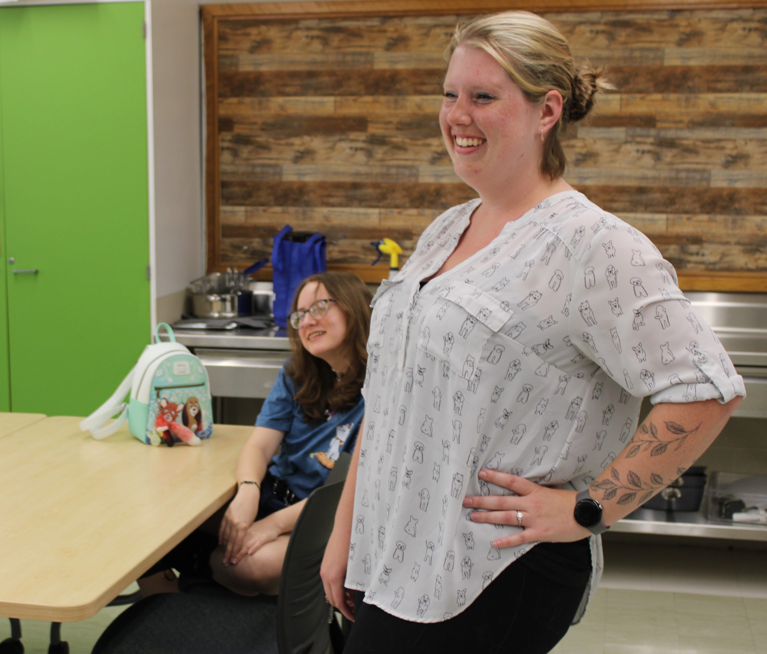 Kasey Mang is pictured in a classroom. She is standing and smiling at a subject not seen in the picture. A student can also be seen seated at a desk, smiling at the same subject as Kasey.