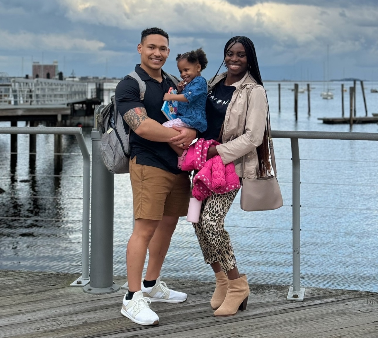 Ryan Kamanu and his wife Vivian pose for a photo with their daughter Sophie. They are standing on a pier. You can see water behind them and it appears to be an overcast day.