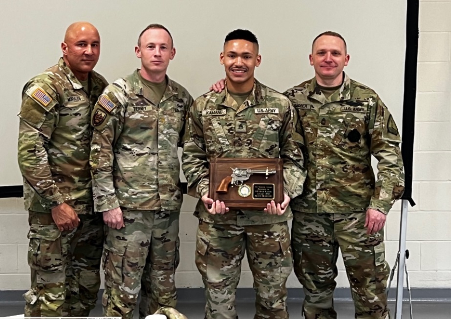 A group of Army National guard members pose for a picture . They are wearing camouflage fatigues. Ryan Kamanu, a PTECH completer is second from the right and is holding an award he received.